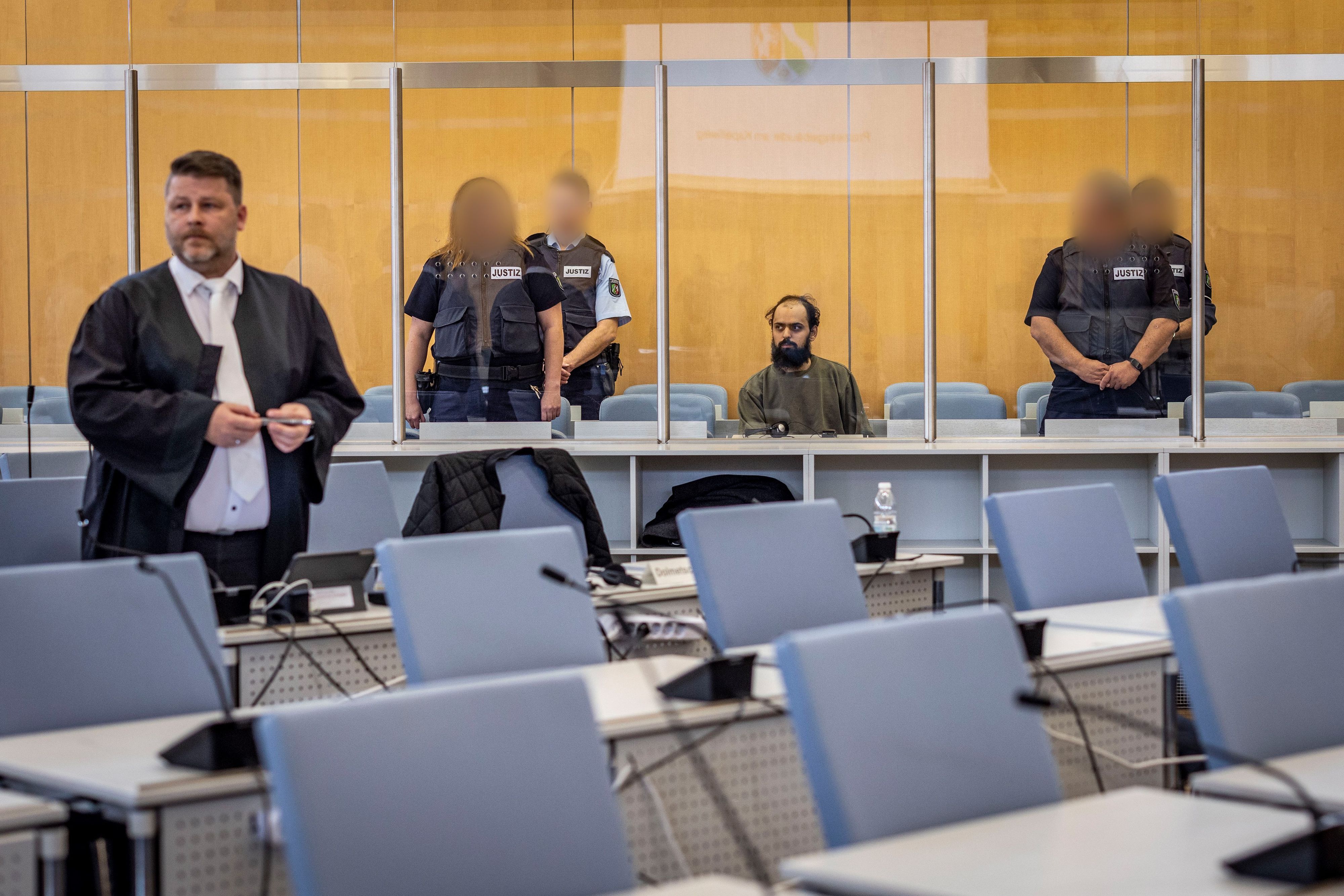 23.10.2023, Nordrhein-Westfalen, Düsseldorf: Der Angeklagte Maan D. (M) wird von Justizbeamten bewacht, er sitzt hinter einer Sicherheitsscheibe auf einem Stuhl. Links steht sein Anwalt Christian Grotenhöfer. ACHTUNG: Justizbeamte wurden auf Anweisung des Gerichts gepixelt +++ dpa-Bildfunk +++