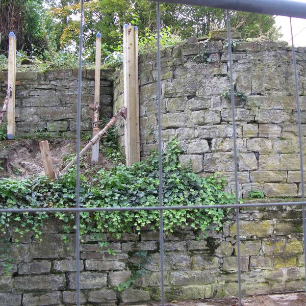 Im Vordergrund steht ein Bauzaun. Dahinter ist die halbrunde Apsis der zerstörten Synagoge zu sehen. Die Bruchsteinmauern sind durch Stützen gesichert.