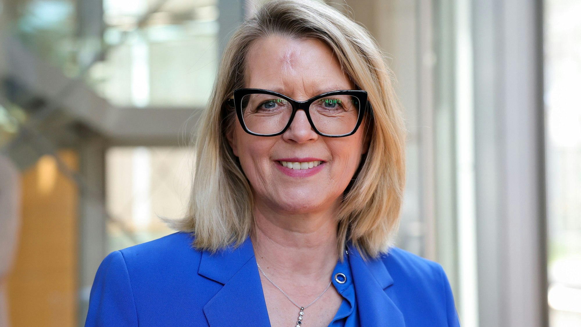 Eine blonde Frau mit Brille und blauem Blazer lächelt in die Kamera.