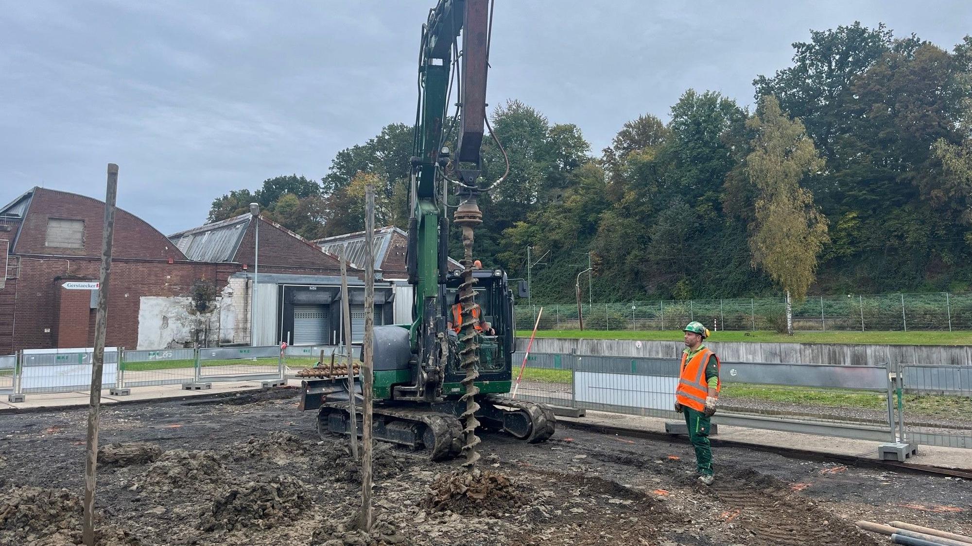 Ein Bagger vor Werkshallen mit einem Sheddach. Seitlich steht ein Arbeiter in roter Sicherheitsweste.