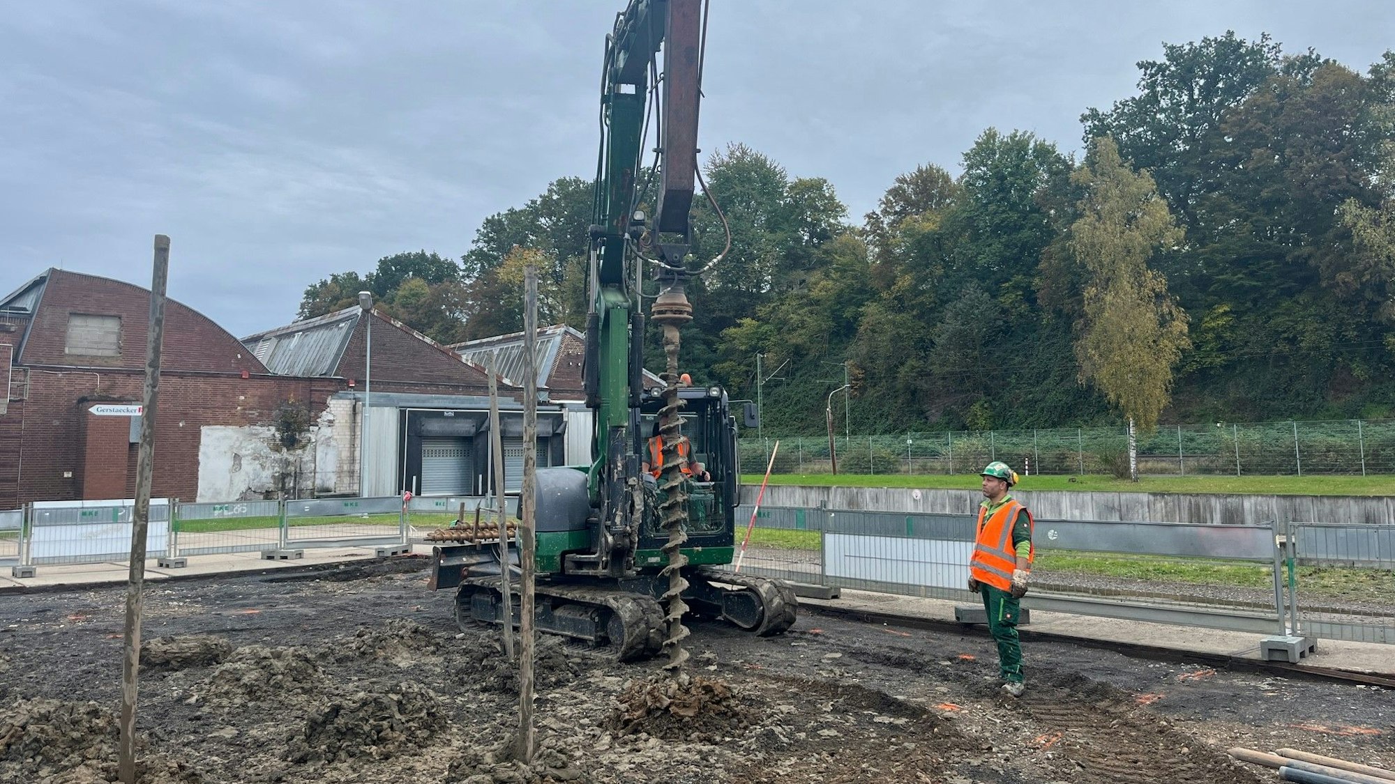 Ein Bagger vor Werkshallen mit einem Sheddach. Seitlich steht ein Arbeiter in roter Sicherheitsweste.