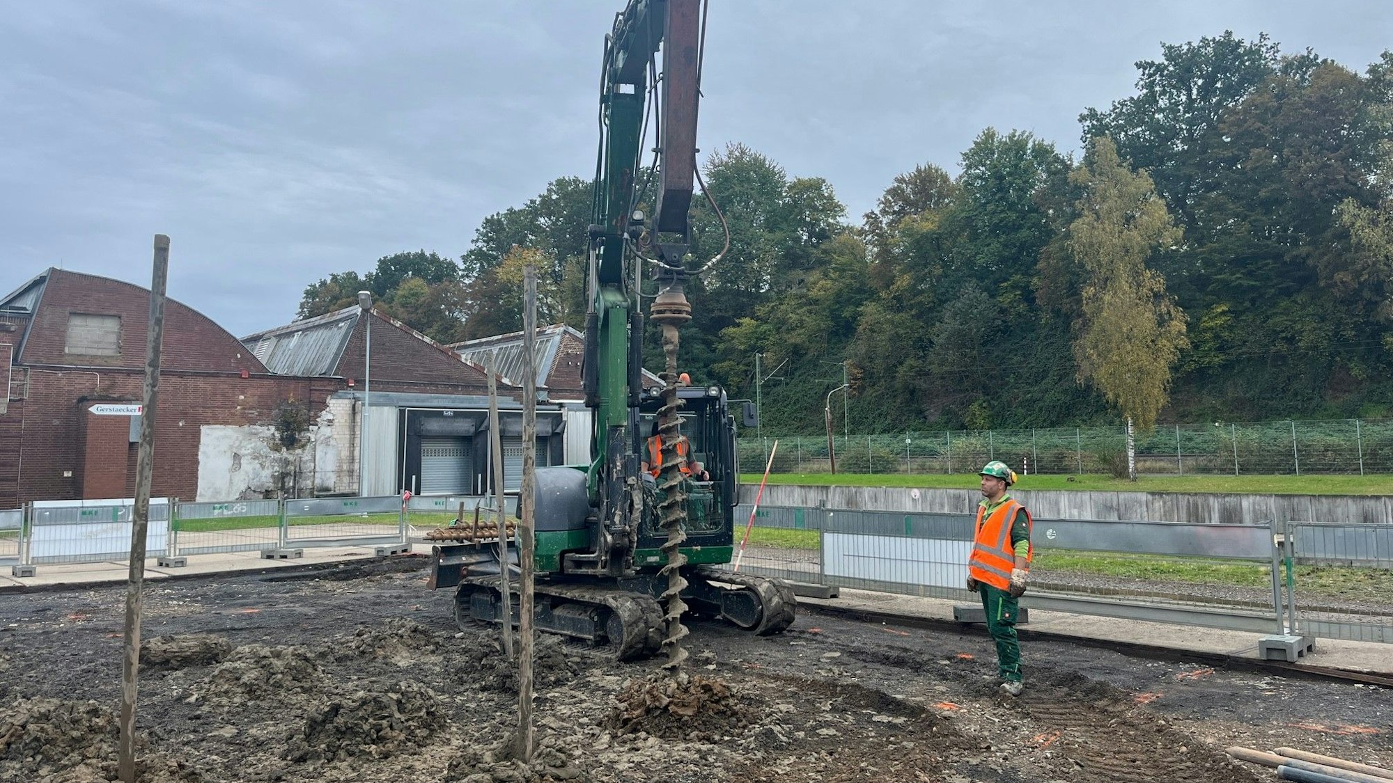 Ein Bagger vor Werkshallen mit einem Sheddach. Seitlich steht ein Arbeiter in roter Sicherheitsweste.