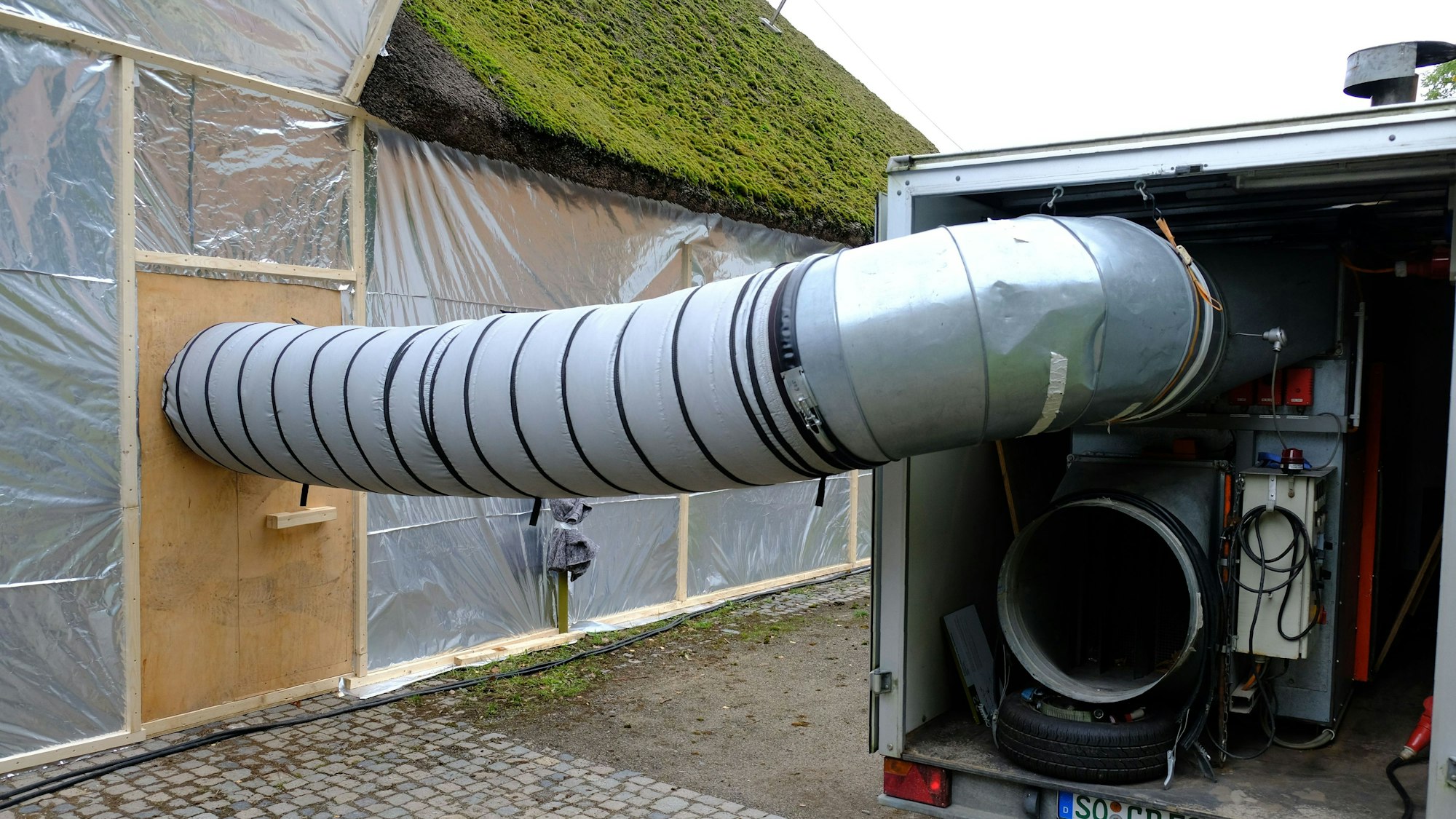 Auf einem Anhänger steht ein Ölbrenner. Von dort führt ein voluminöser Schlauch in eine mit einer silbernen Folie „verpackte“ Fachwerkscheune.