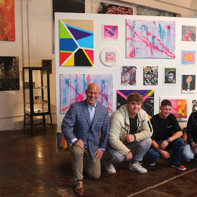 Zwei Männer, eine Frau und zwei Jugendliche knien vor einer Wand mit Gemälden.