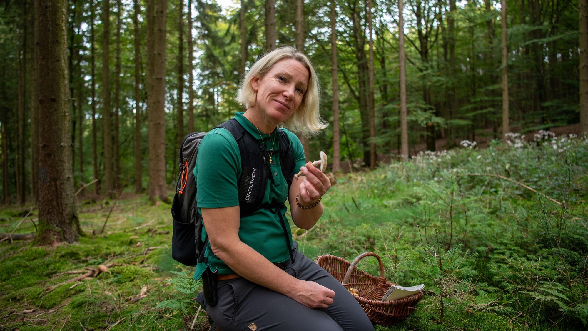 Christina Wojtowicz kniet auf dem Waldboden und hält einen Pilz in der Hand. Neben ihr steht ein Korb, in dem bereits gesammelte Pilze und ein Bestimmungsbuch liegen.