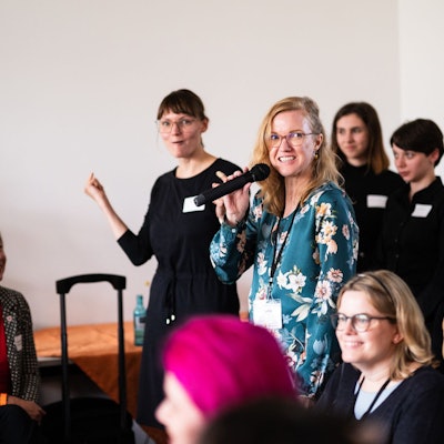 Eine junge blonde Frau mit Mikrofon steht in einer Gruppe Menschen.