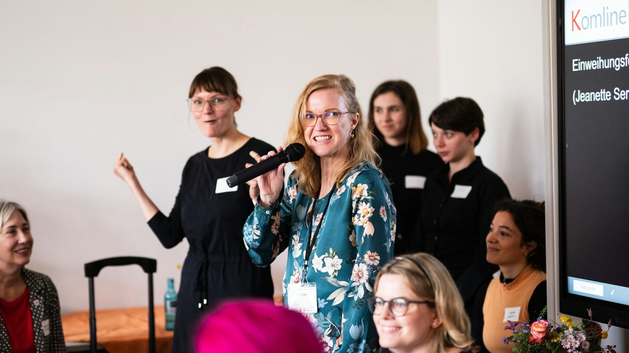 Eine junge blonde Frau mit Mikrofon steht in einer Gruppe Menschen.