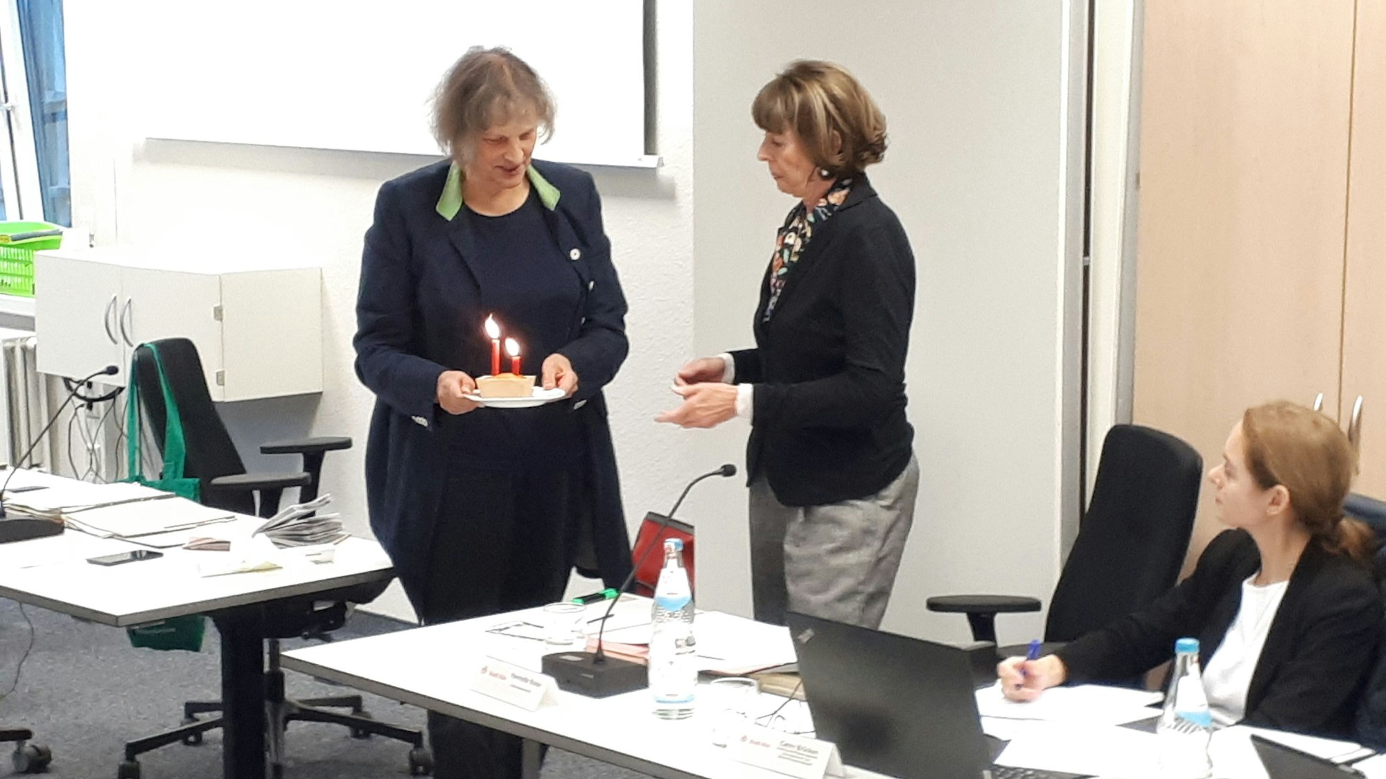Eine Frau übergibt einer anderen ein kleines Törtchen mit zwei brennenden Kerzen darauf.