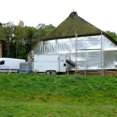 Bis auf das mit Reet gedeckte Dach ist ein großes Scheunengebäude im Freilichtmuseum Kommern in eine silbern glänzenden Folie eingepackt. Aus einem davor stehenden Anhänger führt ein voluminöser Schlauch ins Innere des Gebäudes.