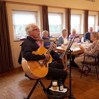 Der Vorsitzende des Vokalensembles, Günter Pieler, sitzt mit der Gitarre auf einem Hocker, vor ihm das Publikum mit Textblättern in der Hand.
