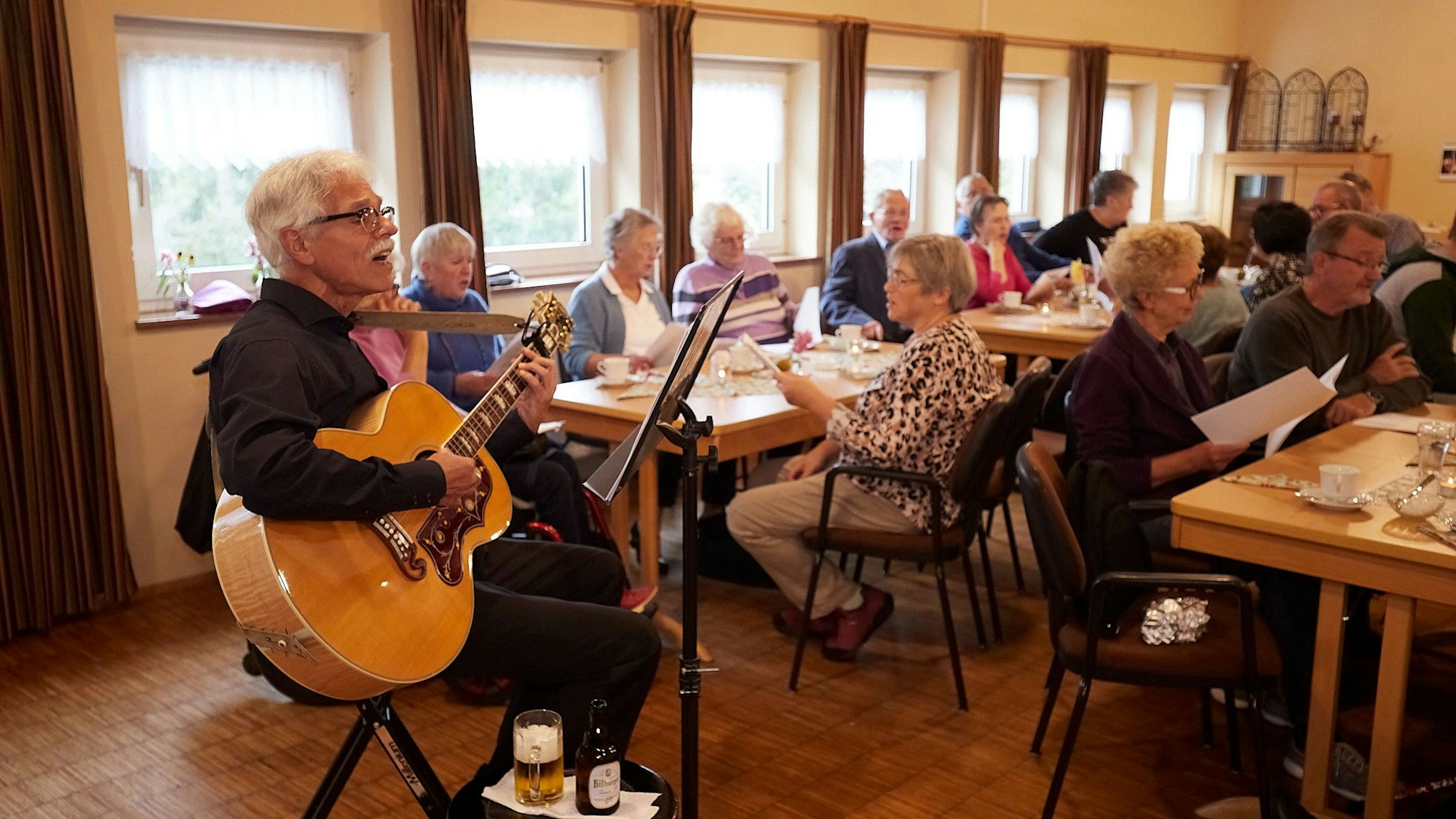 Der Vorsitzende des Vokalensembles, Günter Pieler, sitzt mit der Gitarre auf einem Hocker, vor ihm das Publikum mit Textblättern in der Hand.