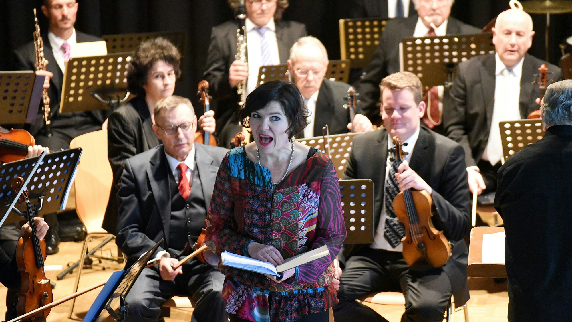 Eine Frau rezitiert vor einem Orchester stehend.