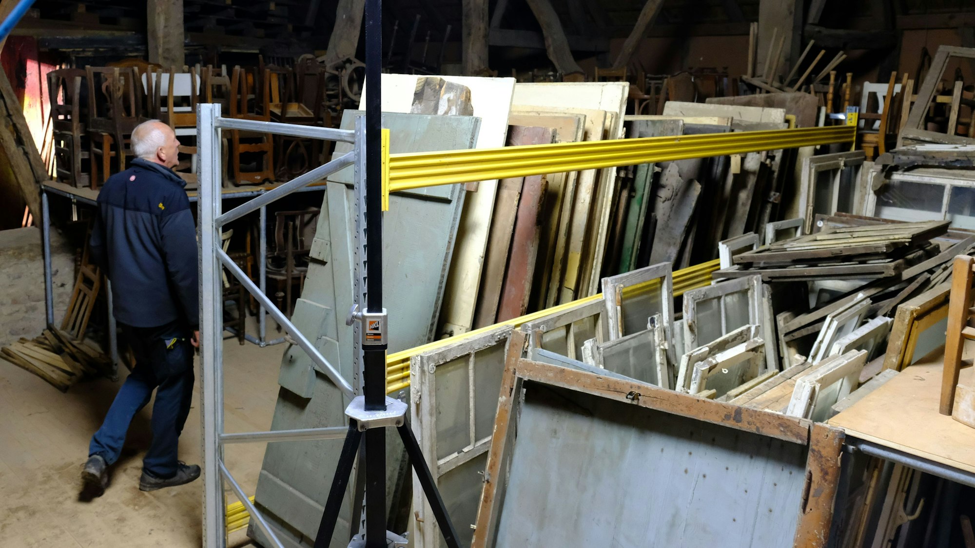 Ein Mann in Arbeitskleidung geht durch eine Scheune, in der sich zahlreiche Möbel und Holzbauteile befinden.