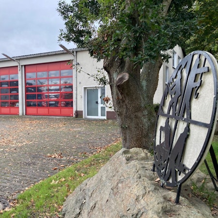 Das Feuerwehrgerätehaus in Habbelrath ist von außen zu sehen, rechts das Logo der Feuerwehr, daneben ein Baum.