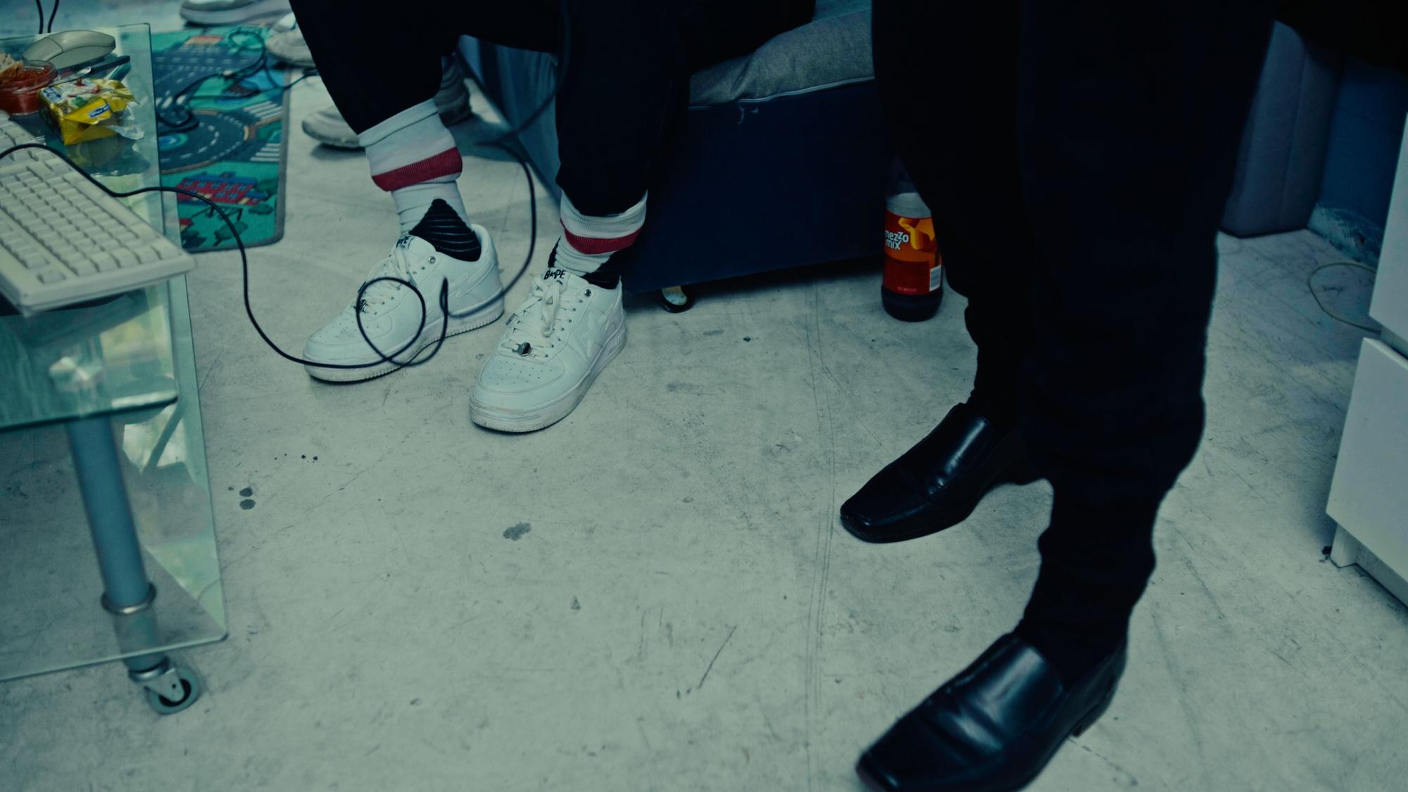 Das Bild zeigt die Schuhe von Equal und Liaze. Equal trägt weiße Sneaker und Tennissocken, in denen seine Hose steckt. Liazes Füße stecken in schwarzen Lederslippern, die er zur Jogginghose trägt.