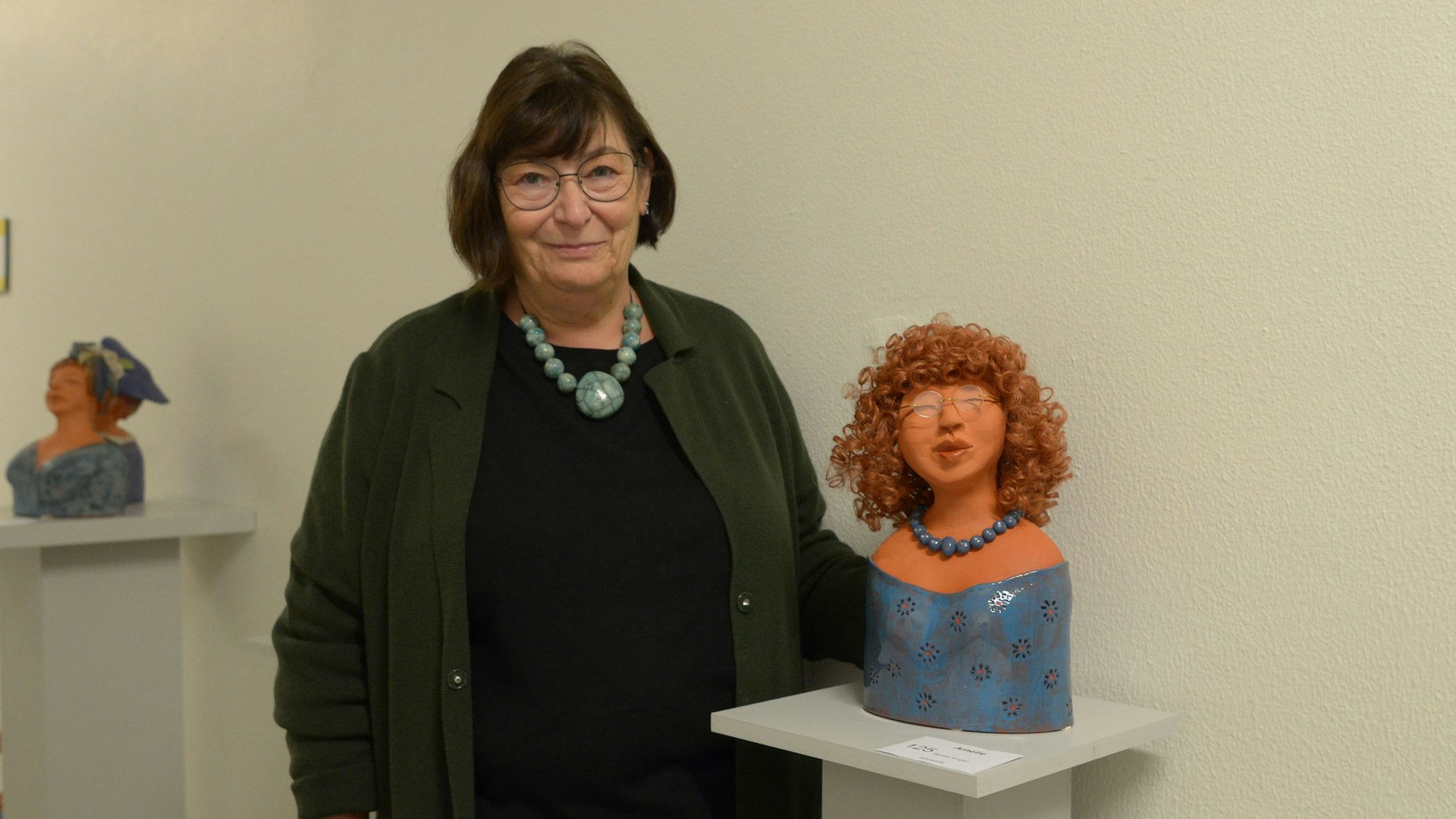 Die Künstlerin Beate Krupp steht neben einer ihrer Keramikskulpturen. Es handelt sich um eine Frauenfigur mit Perücke, Brille und Halskette.