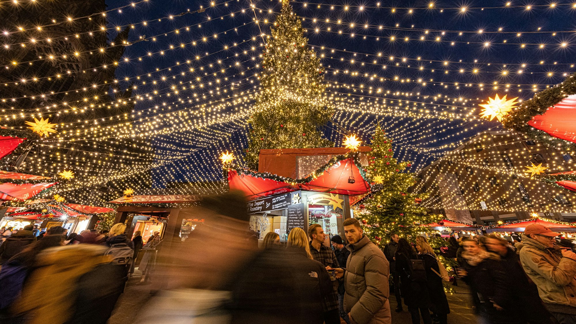 Leute stehen an den Buden mit roten Dächern. Lichterketten bilden einen Sternenhimmel.