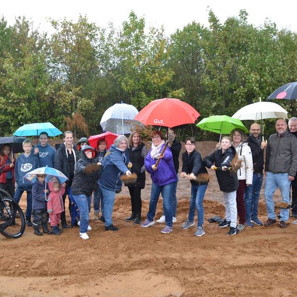 Eine größere Gruppe Menschen, Kinder und Erwachsene, steht mit Regenschirmen auf einem Bauplatz. Einige haben mit einem Spaten einen symbolischen Spatenstich vollzogen.