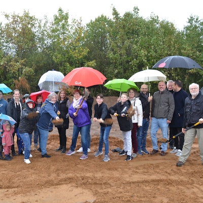 Eine größere Gruppe Menschen, Kinder und Erwachsene, steht mit Regenschirmen auf einem Bauplatz. Einige haben mit einem Spaten einen symbolischen Spatenstich vollzogen.