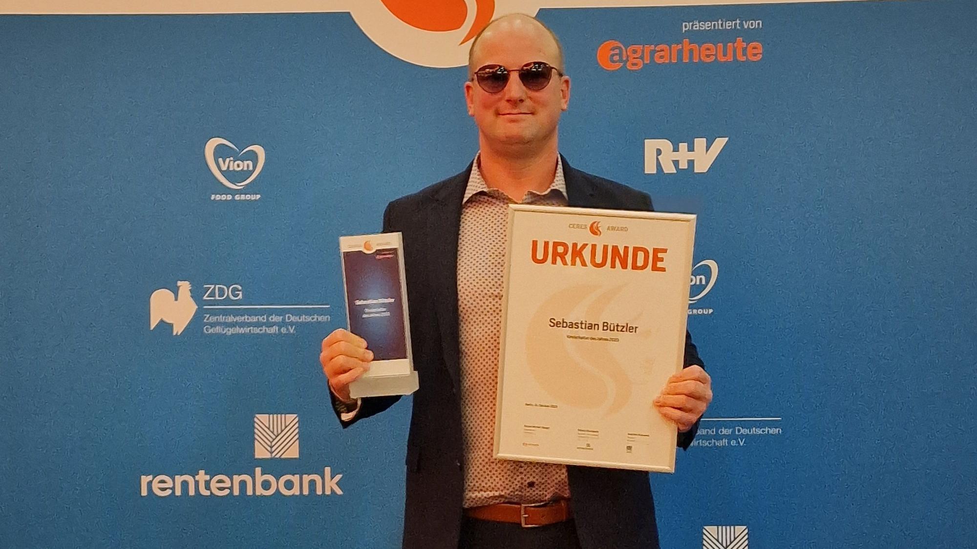 Sebastian Bützler steht vor einem Plakat des Ceres-Awards und hält einen Glas-Pokal sowie eine Urkunde in der Hand.