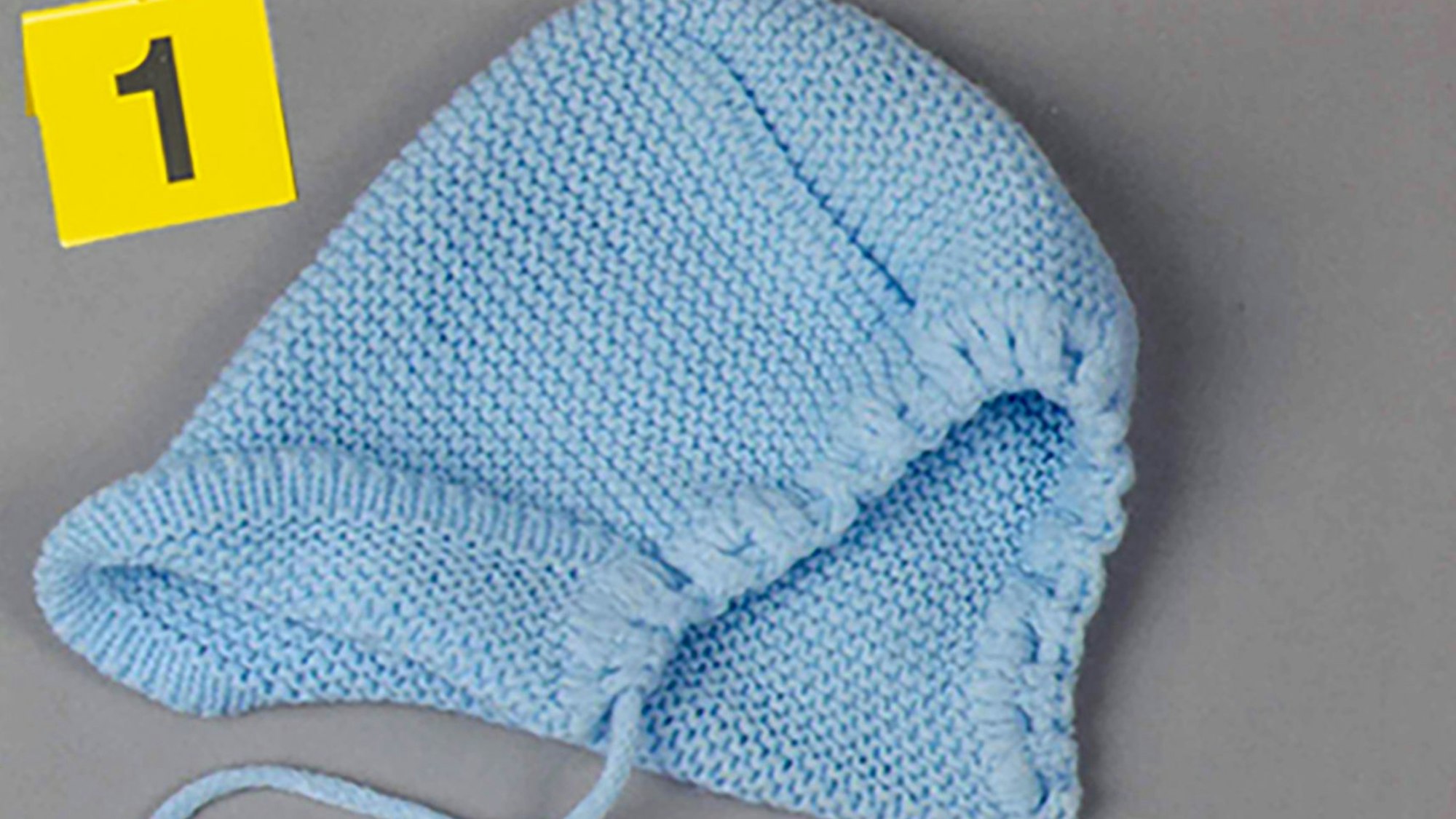 Eine hellblaue Babymütze liegt auf einem Tisch. Darüber steht ein gelbes Schild mit der Nummer 1.