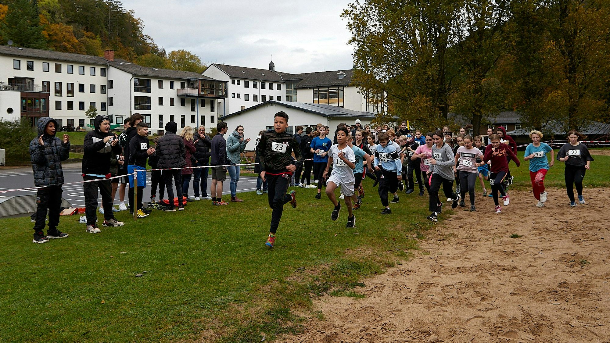 Zahlreiche junge Läufer starten zu einem Crosslauf. Hinter einem Flatterband stehen Jugendliche und Erwachsene und feuern sie an.