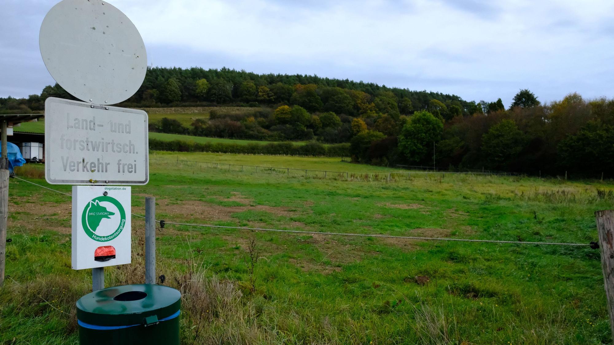 Eine Wiese in Hanglage am Ortsrand von Kallmuth. Links sind ein verblichenes Verkehrsschild, ein Spender für Hundekottüten und ein Mülleimer zu sehen.