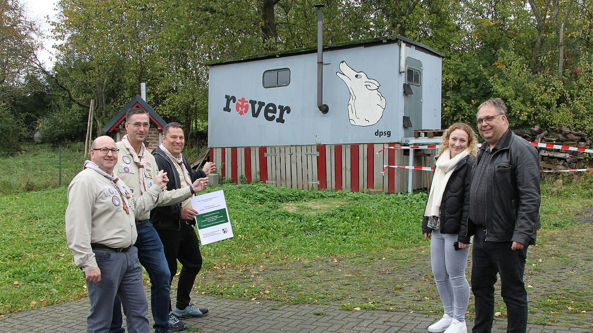 Vier Männer und eine Frau stehen vor einem alten, grauen Bauwagen, der mit einem Schriftzug „rover“ und dem weißen Wolf als Zeichen des Pfadfinderstammes Rohr-Lindweiler gestaltet ist.