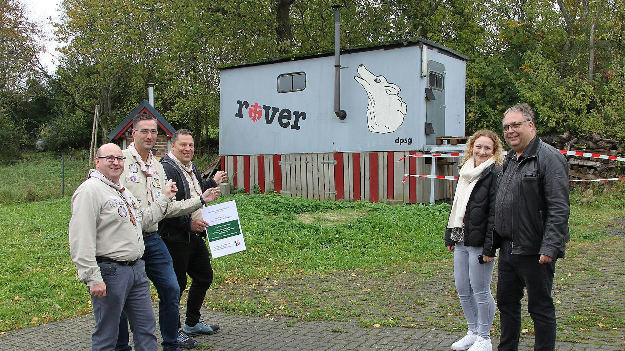 Vier Männer und eine Frau stehen vor einem alten, grauen Bauwagen, der mit einem Schriftzug „rover“ und dem weißen Wolf als Zeichen des Pfadfinderstammes Rohr-Lindweiler gestaltet ist.