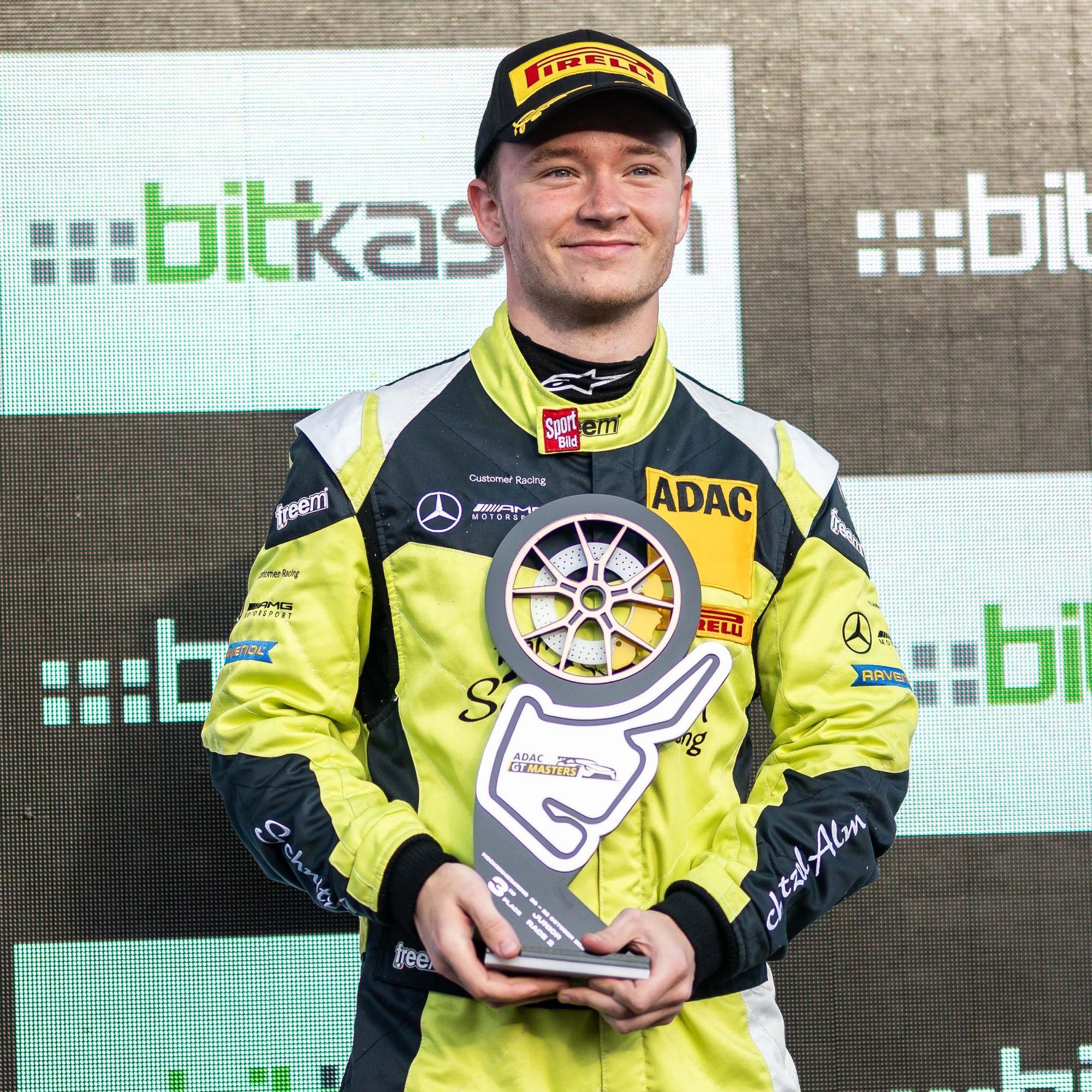 Moritz Wiskirchen steht in einem Rennanzug vor einer Sponsoren-Wand und hält einen Pokal in den Händen.
