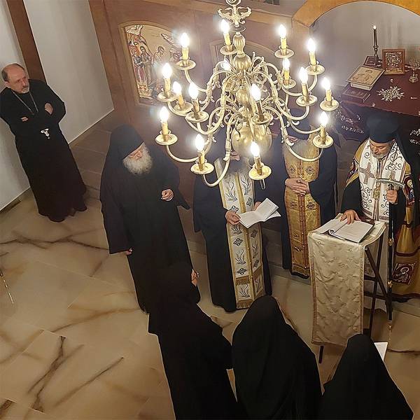 Orthodoxe Priester und Nonnen stehen in einer Kapelle unter einem Kronleuchter. An den holzvertäfelten Wänden sind sakrale Gemälde zu sehen.