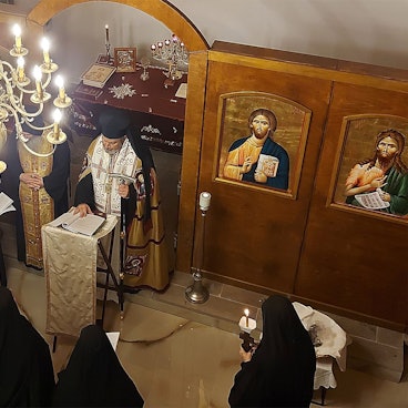 Orthodoxe Priester und Nonnen stehen in einer Kapelle unter einem Kronleuchter. An den holzvertäfelten Wänden sind sakrale Gemälde zu sehen.