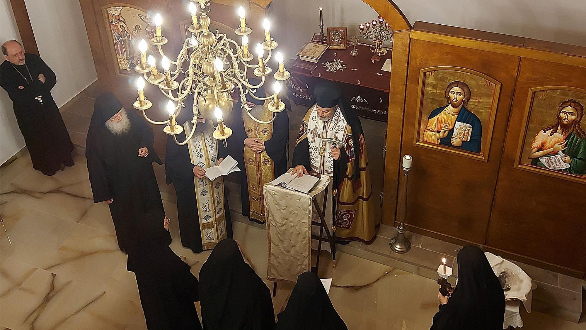 Orthodoxe Priester und Nonnen stehen in einer Kapelle unter einem Kronleuchter. An den holzvertäfelten Wänden sind sakrale Gemälde zu sehen.