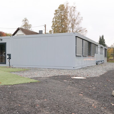 Vor einer Container-Unterkunft stehen leere Plastikstühle auf Kunstrasenteppichen.