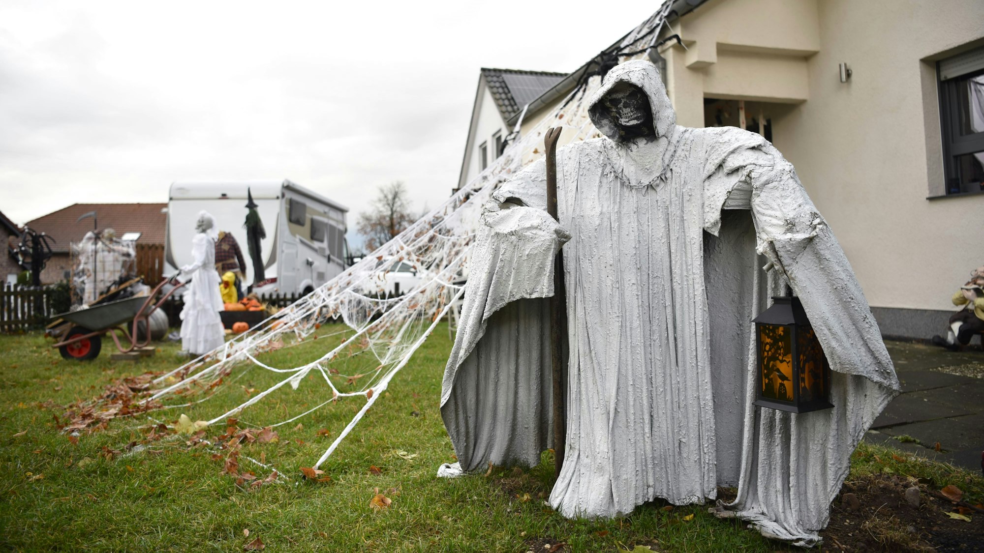Eine Figur in einem langen weißen Mantel, mit Kapuze, Sense und Laterne steht vor einem Haus. Im Hintergrund sind künstliche Spinnweben, Skelette und Kürbisse zu erkennen.