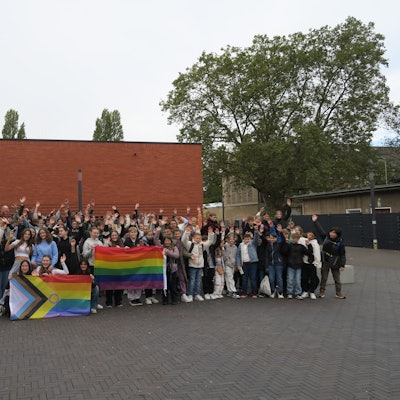 Eine Menge von Schülerinnen und Schülern hat sich auf einem Schulhof zum Foto aufgestellt, sie haben zwei bunte Flaggen bei sich.