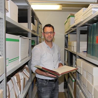 Der Dahlemer Kämmerer Frank Hütter steht zwischen zwei hohen Regalen im Gemeindearchiv. Er hält eine aufgeschlagene Akte in der Hand.