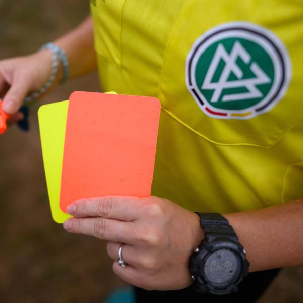 Eine Schiedsrichterin steht mit roter und gelber Karte auf einem Rasen und hält in der anderen Hand eine Trillerpfeife.