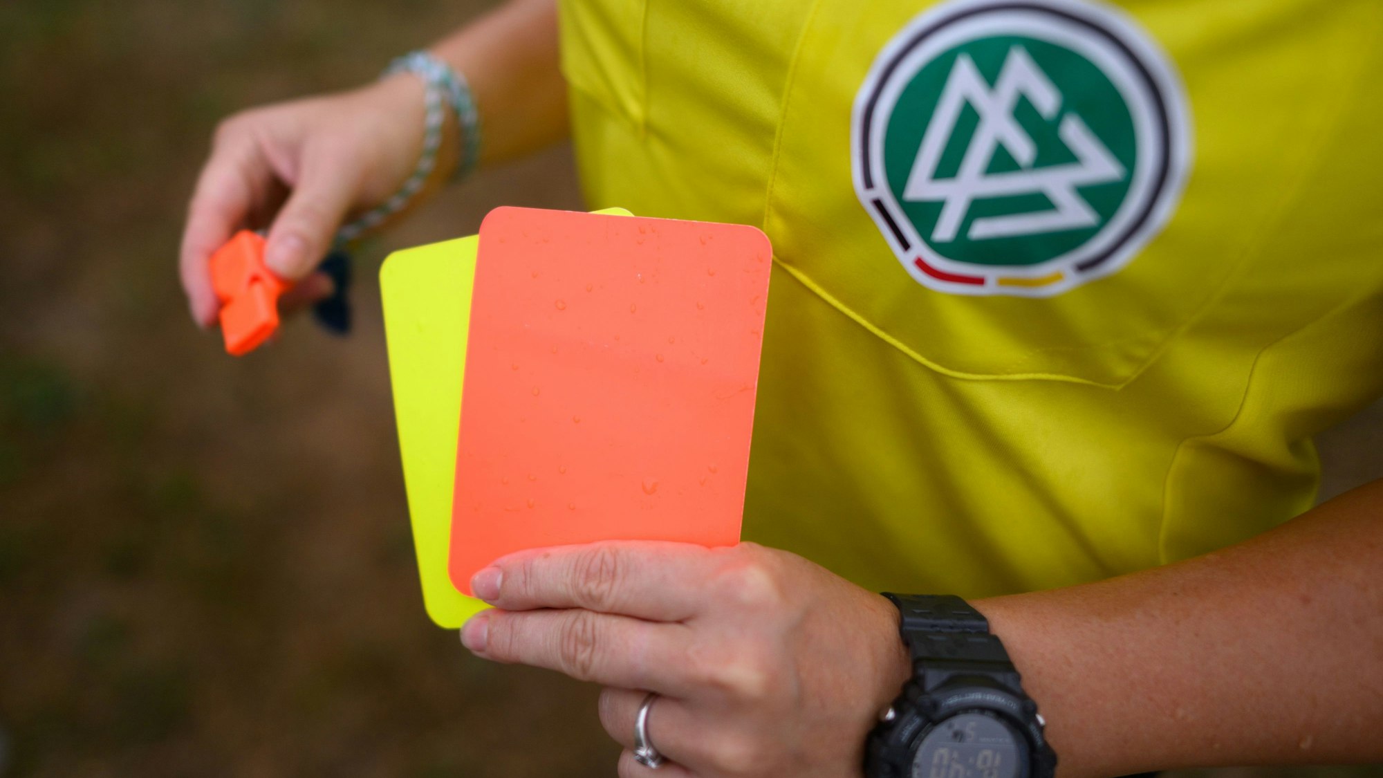 Eine Schiedsrichterin steht mit roter und gelber Karte auf einem Rasen und hält in der anderen Hand eine Trillerpfeife.