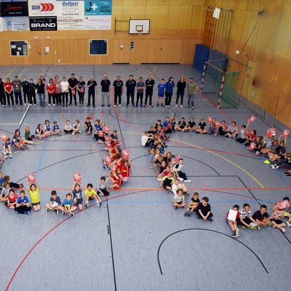 Zahlreiche Sportlerinnen und Sportlern sitzen in einer Halle auf dem Boden und bilden gemeinsam eine 100.