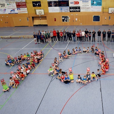 Zahlreiche Sportlerinnen und Sportlern sitzen in einer Halle auf dem Boden und bilden gemeinsam eine 100.