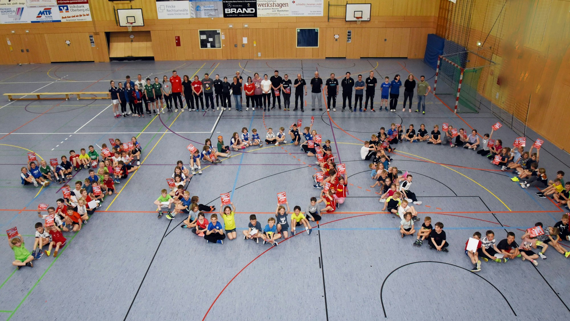 Zahlreiche Sportlerinnen und Sportlern sitzen in einer Halle auf dem Boden und bilden gemeinsam eine 100.