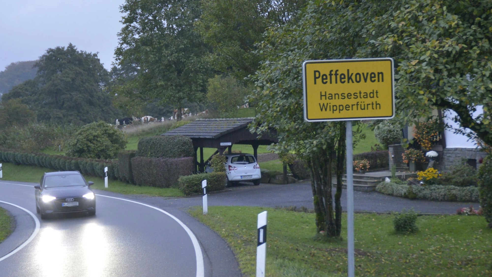 Das Ortsschild von Wipperfürth-Peffekoven steht am Rande einer Straße, ein Auto fährt mit eingeschalteten Scheinwerfern daran vorbei.