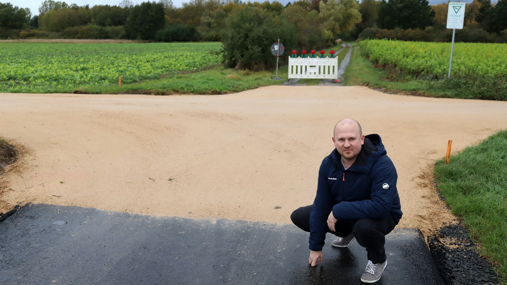 CDU-Ratsherr Martin Finder hockt auf dem geteerten Stück des Radwegs. Dahinter, an der Grenze zum Naturschutzgebiet, hat der Radweg eine wassergebundene Deckschicht.