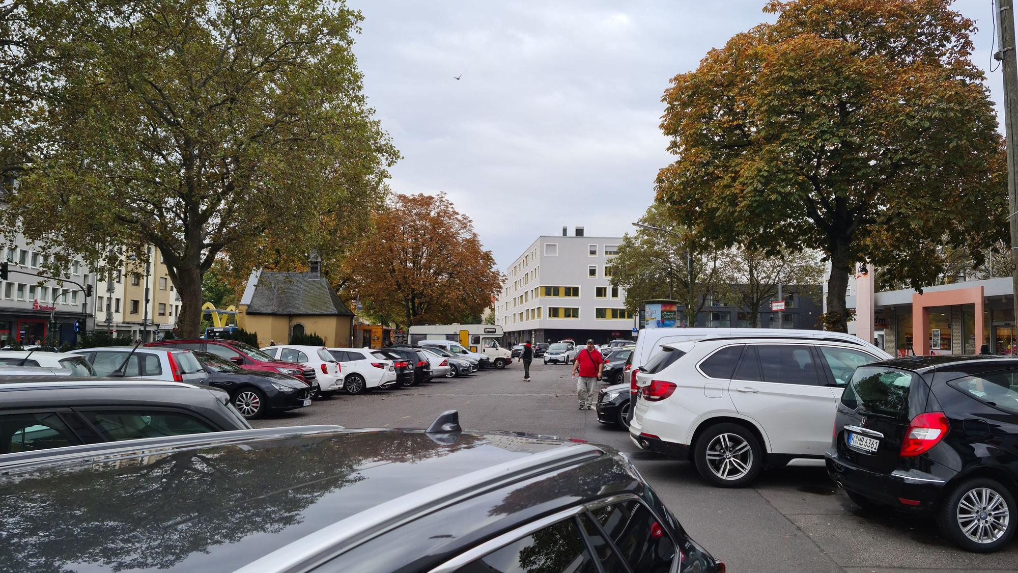 Ein langgezogener Platz mit vielen parkenden Autos und einigen Bäumen am Rand ist zu sehen. Auch eine kleine Kapelle steht am linken Rand der Platzfläche.