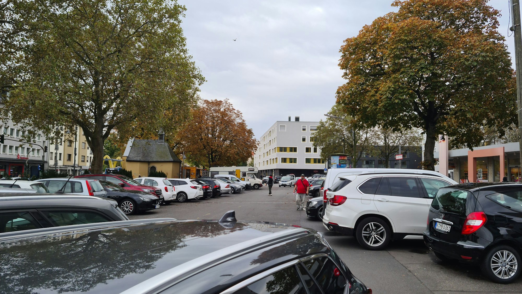 Ein langgezogener Platz mit vielen parkenden Autos und einigen Bäumen am Rand ist zu sehen. Auch eine kleine Kapelle steht am linken Rand der Platzfläche.