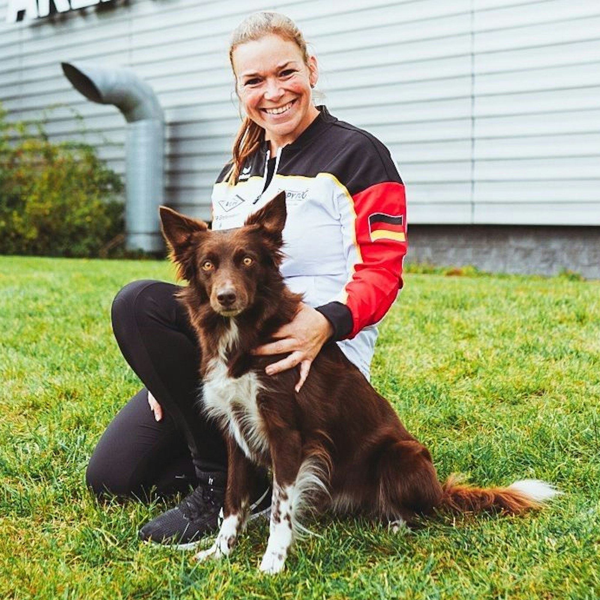 Mona Grefenstein kniet mit ihrer Border-Collie-Hündin Sea auf einer Wiese vor einer Sport-Arena.