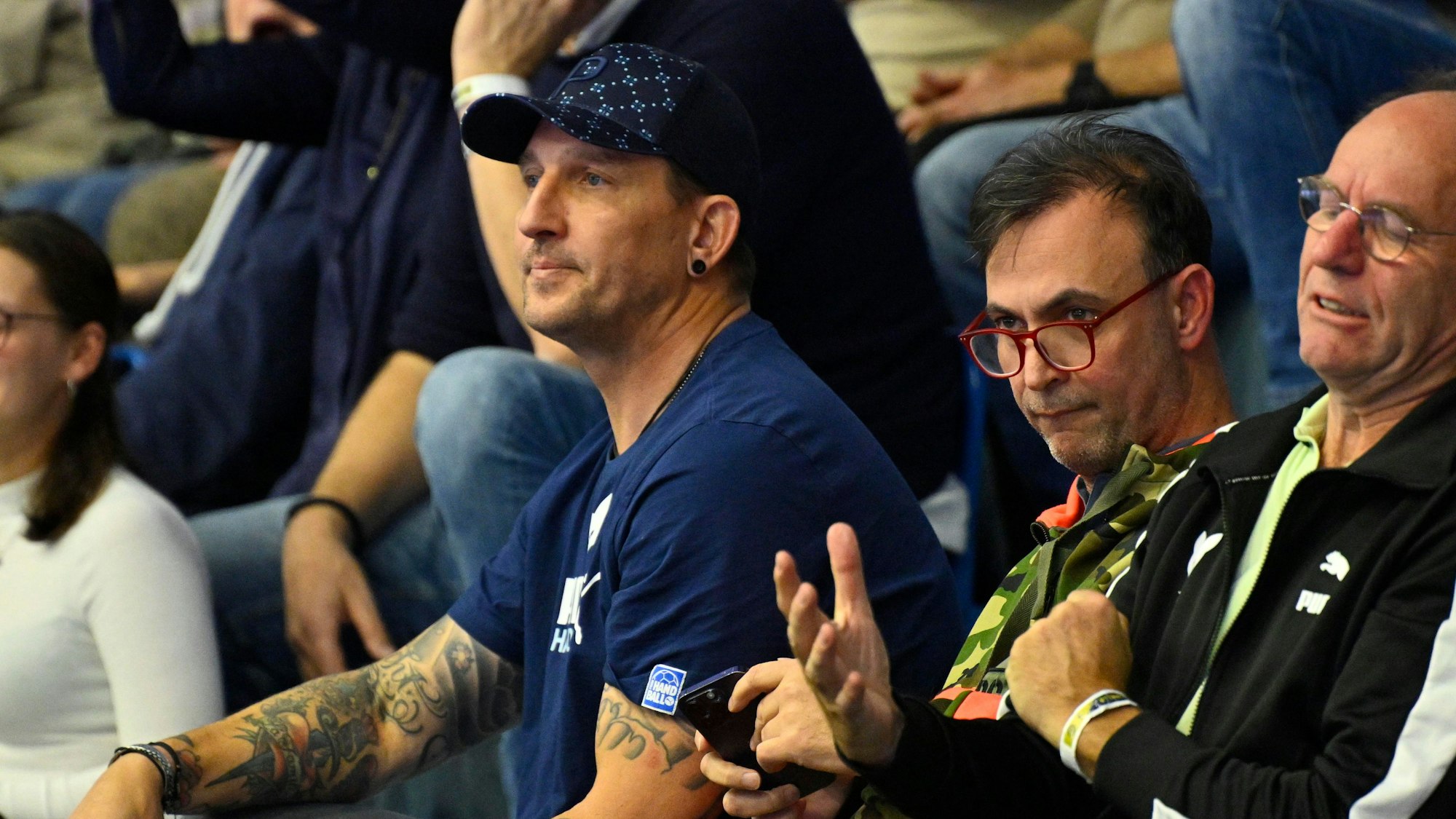 Mehrere Männer sitzen im Publikum bei einem Handballspiel.