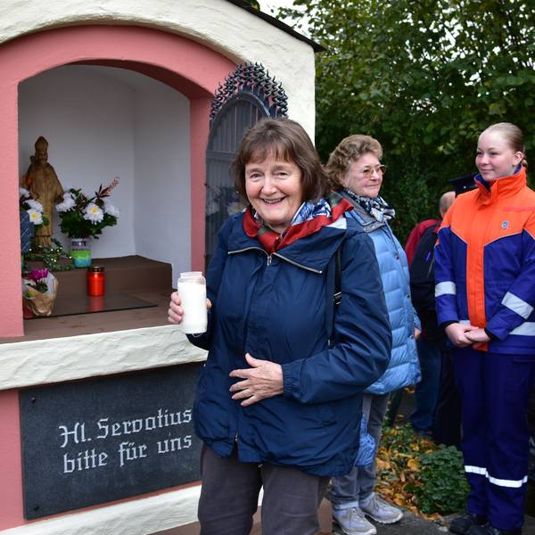 Frau hält Kerze in linker Hand, hinter ihr ein kleines Häuschen mit Blumengestecken und Kerzen