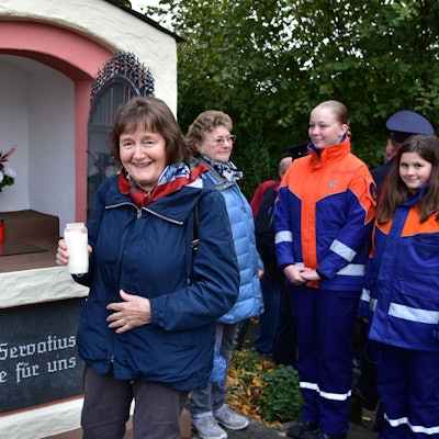 Frau hält Kerze in linker Hand, hinter ihr ein kleines Häuschen mit Blumengestecken und Kerzen